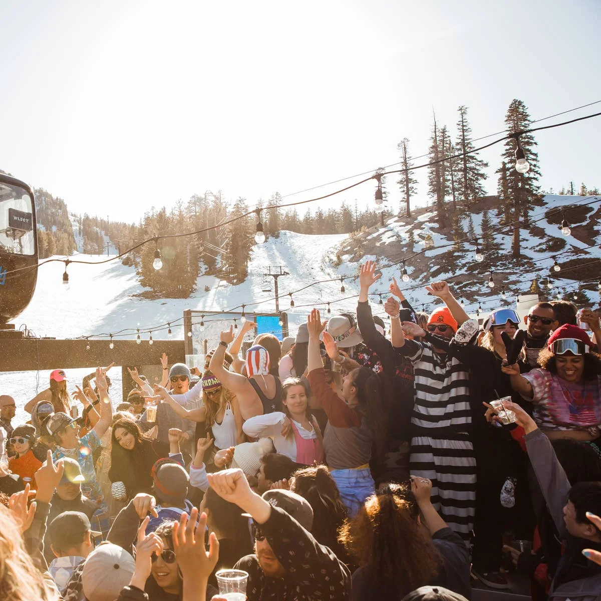 A packed sunny deck at the base of a ski area, a crowd of skiers dancing under string lights with the slopes behind