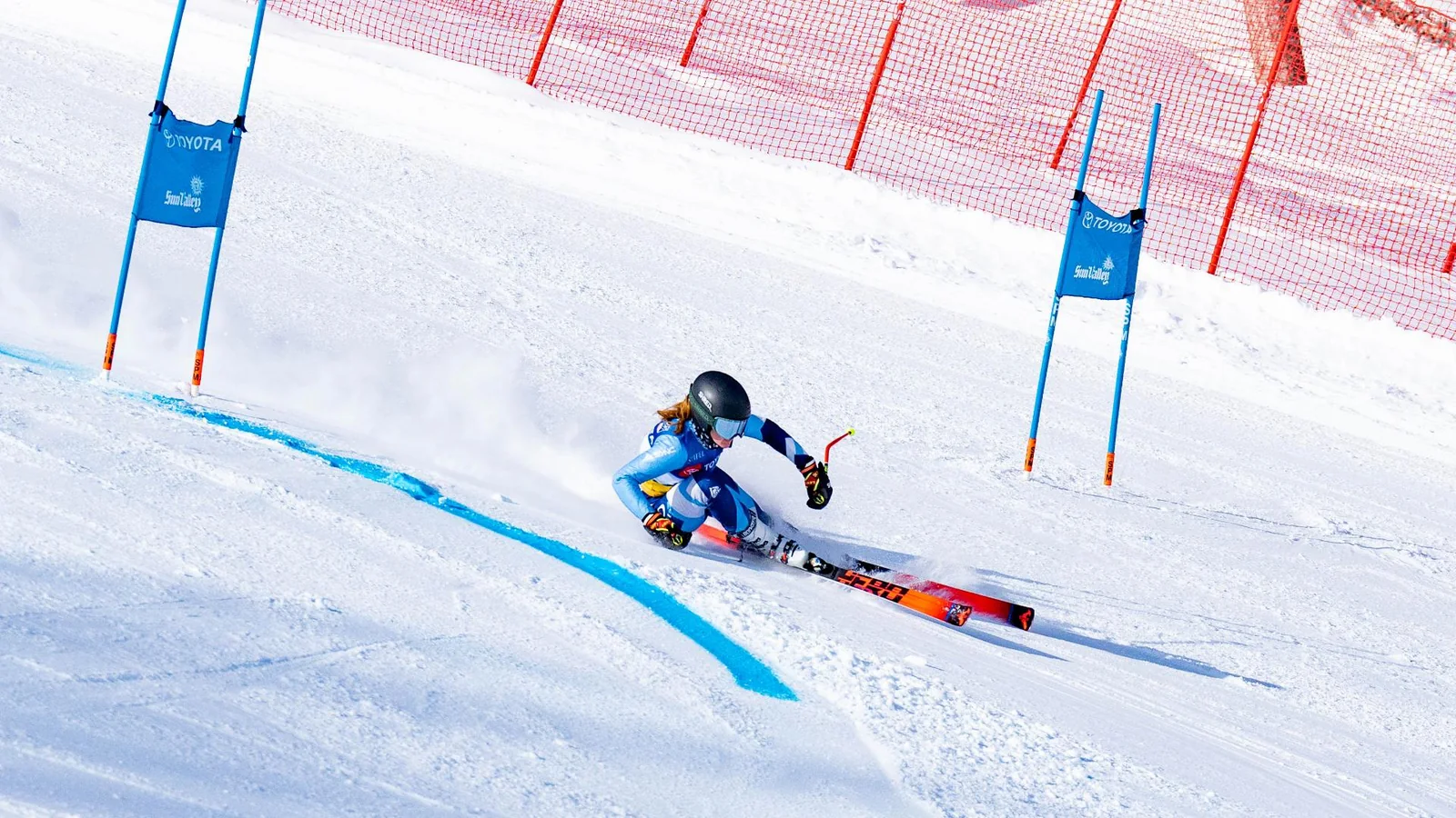 A giant slalom racer carving hard between gates, with orange safety netting behind
