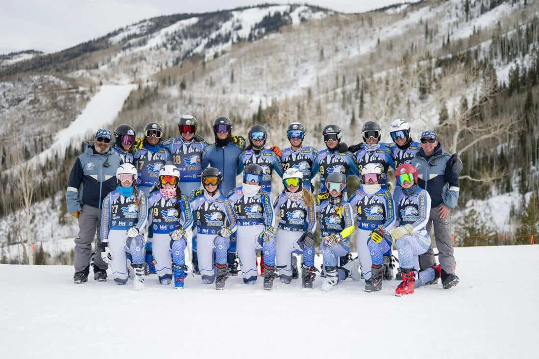 A ski race team in matching race suits lined up with two coaches at the bottom of a race hill