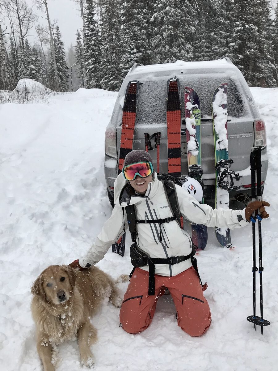 Madie kneeling with their dog beside a ski-loaded truck on a snowy road