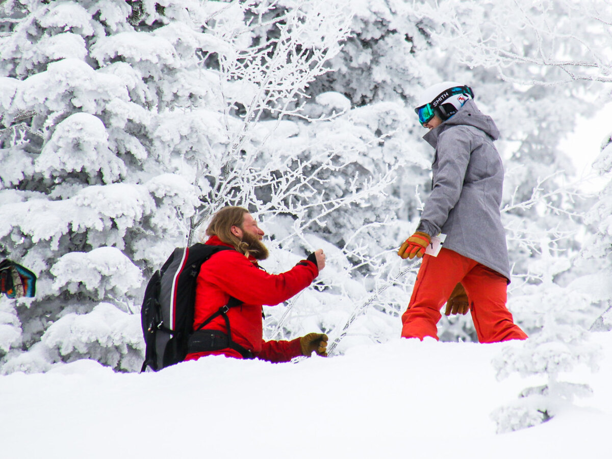 Bass on one knee in deep snow, proposing to Madie among snow-covered trees